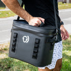 A person outdoors on grass near a road carries The Cave Cooler—a soft cooler bag with a white logo and shoulder strap. Its leak-proof design delivers reliable cold retention for your refreshments. The person wears a black shirt and patterned white shorts.
