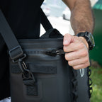 A person wearing a black watch unzips The Cave Cooler, a rugged, leak-proof shoulder bag, outdoors. The focus is on their hand, the zipper, and part of the bag’s strap and handle.