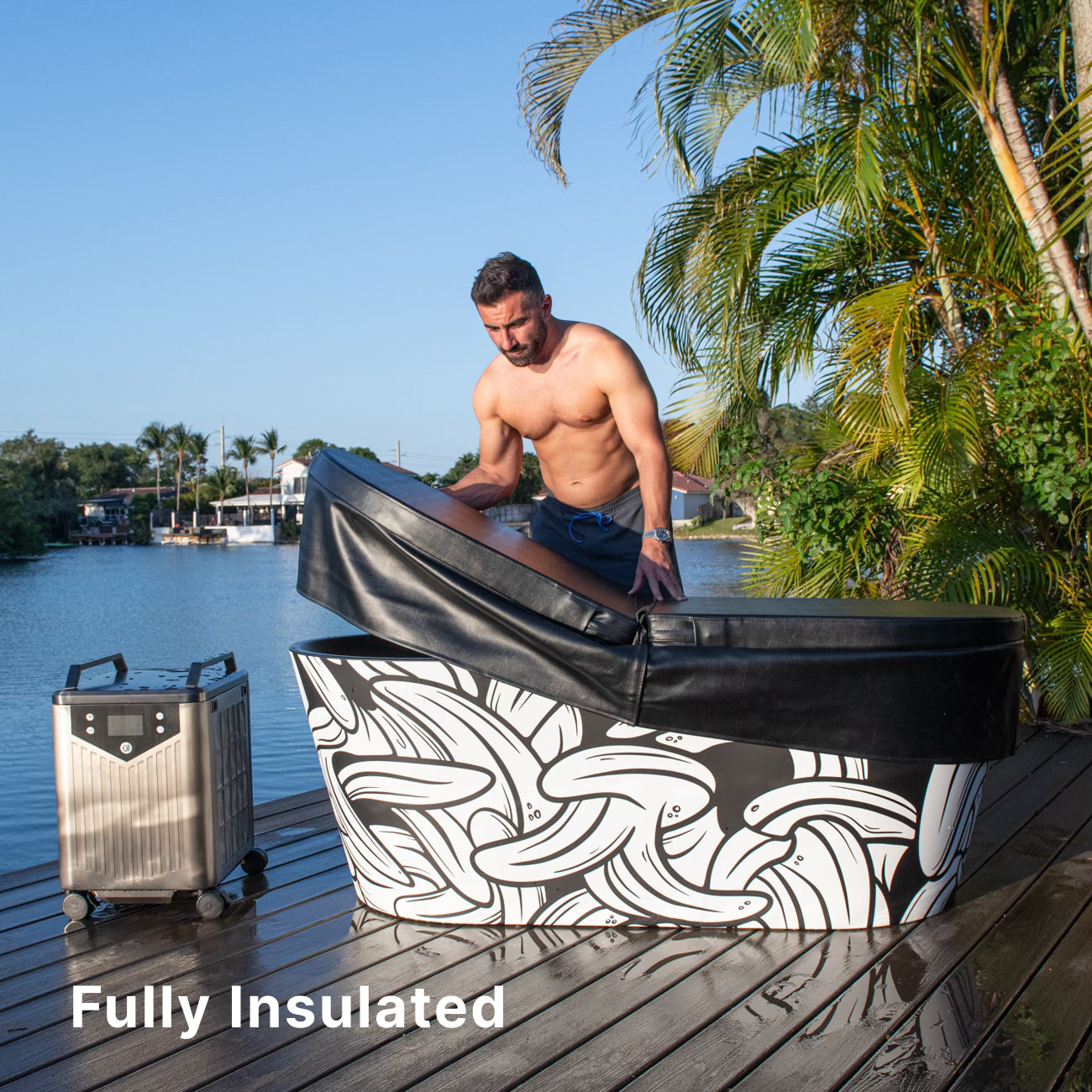 A shirtless man opens the insulated lid of a patterned ice bath tub on a wooden deck by the water, with a portable cooling unit nearby. Palm trees and houses are in the background. Text reads “Fully Insulated.”.