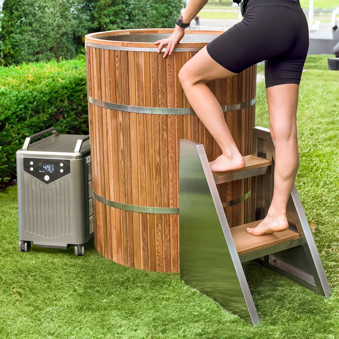 A person in black shorts steps up metal stairs into the Star Treatment 2.0 wooden outdoor cold plunge tub on a grassy lawn. A portable chiller with digital display sits nearby, ideal for compact spaces, while green hedges border the scene.