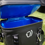 A close-up of The Cave Cooler, an open black soft cooler with a bright blue interior, leak-proof construction, zipper, handles, and a white logo on the front, sitting on green grass.
