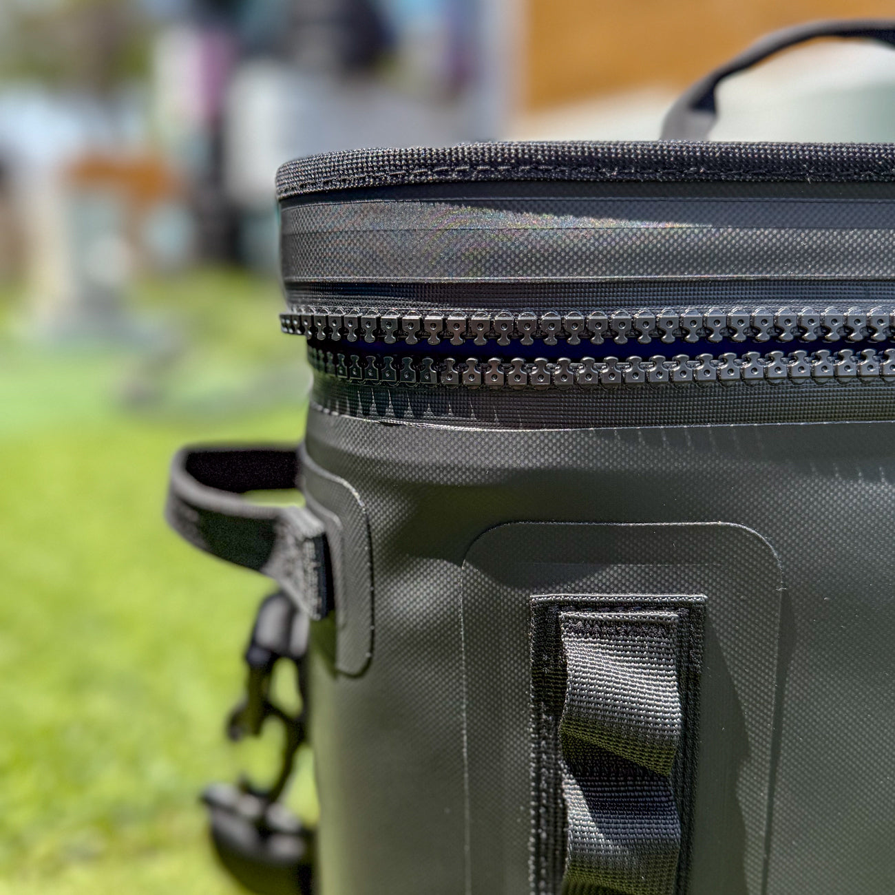 Close-up of The Cave Cooler, a black leakproof soft cooler bag with a handle, displayed outdoors on a sunny day with blurred green grass and buildings in the background.