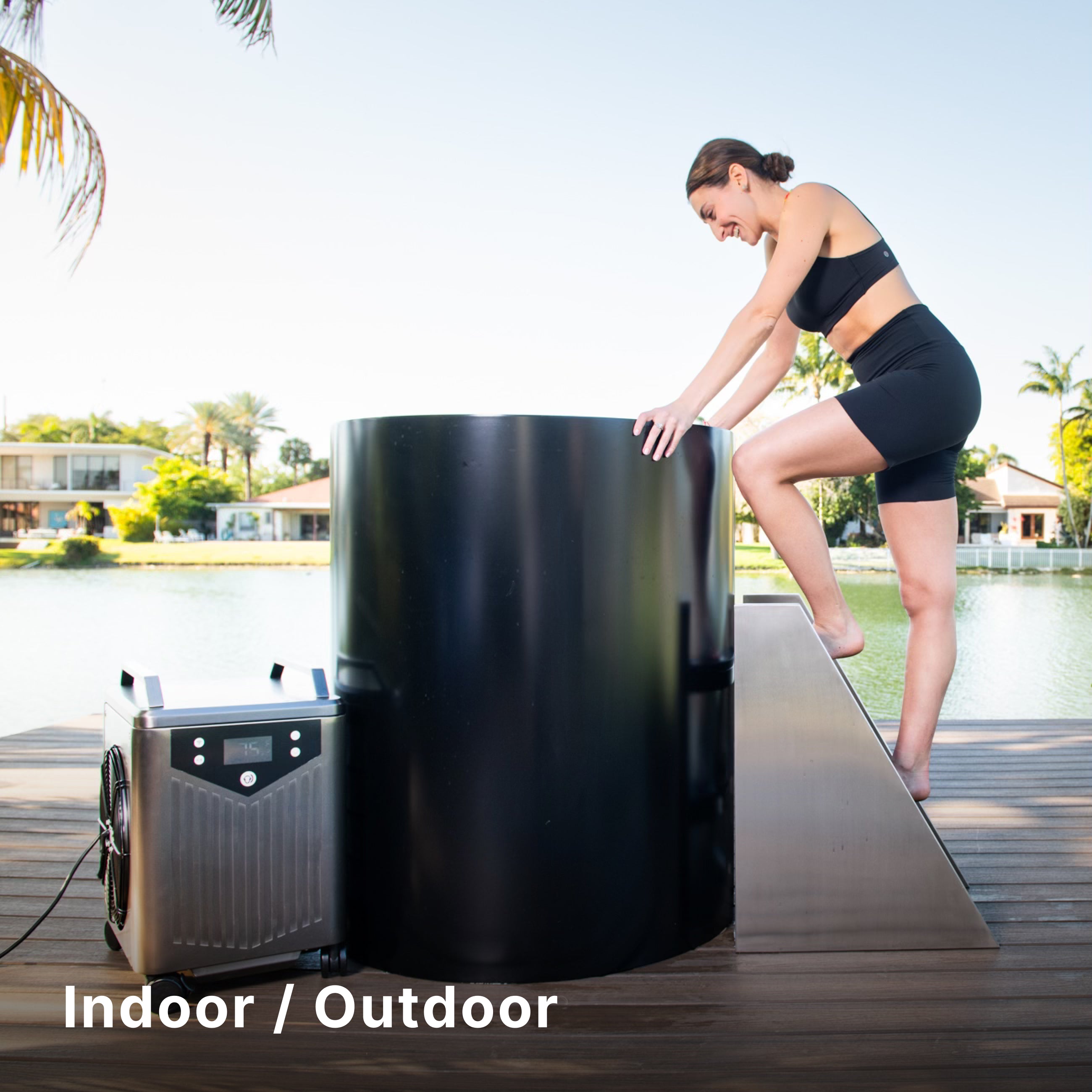 A woman in athletic wear smiles as she climbs into a large black tub next to a portable cooling unit on a wooden deck by the water, with trees and houses in the background. Text reads “Indoor / Outdoor.”.