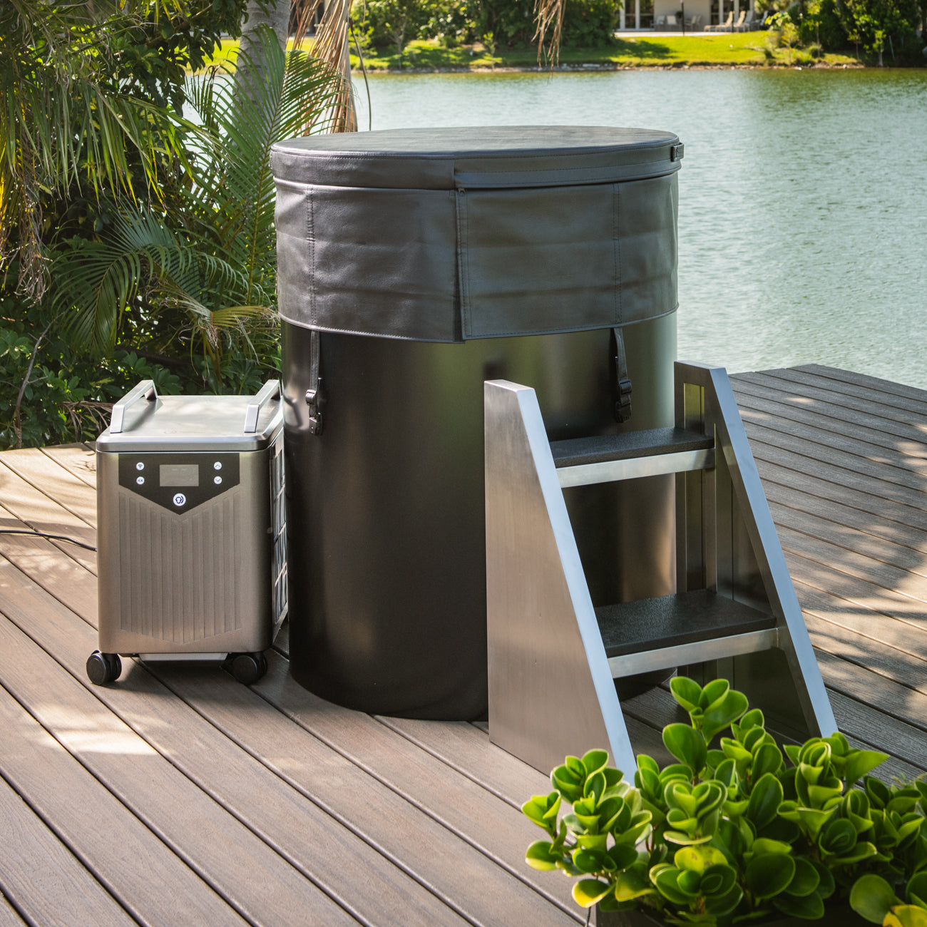 The Cyber Barrel, a black vertical cold plunge tub with a covered lid, sits on a wooden deck by the water beside steps and its portable wheeled control unit. Greenery and trees create a scenic backdrop.