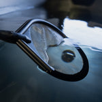 A close-up of the Skimmer Net, a black stainless steel mesh net partially submerged in water, lifting a stingray. The reflective surface highlights its use as a cold plunge skimmer for debris removal, with the stingray visible inside.
