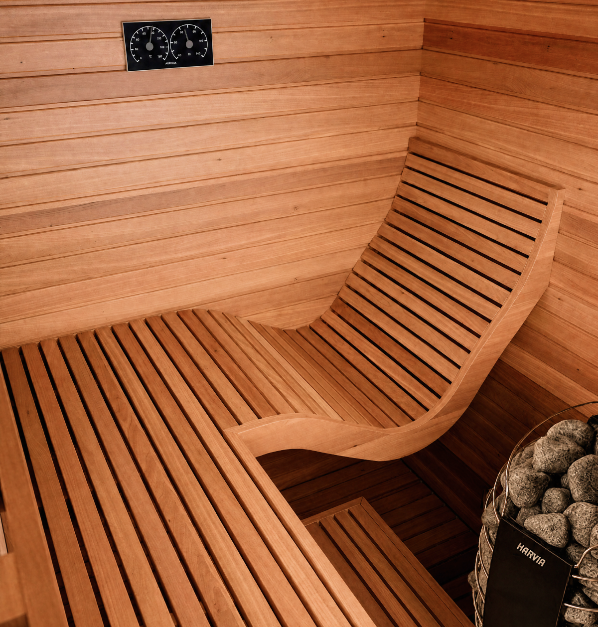 The Baia Glass Sauna features a modern wooden interior with a curved bench, temperature and humidity gauges mounted on the wall, and a Harvia-brand sauna heater with stones situated in the corner. An elegant glass sauna door enhances the sleek space, creating an open and inviting atmosphere.