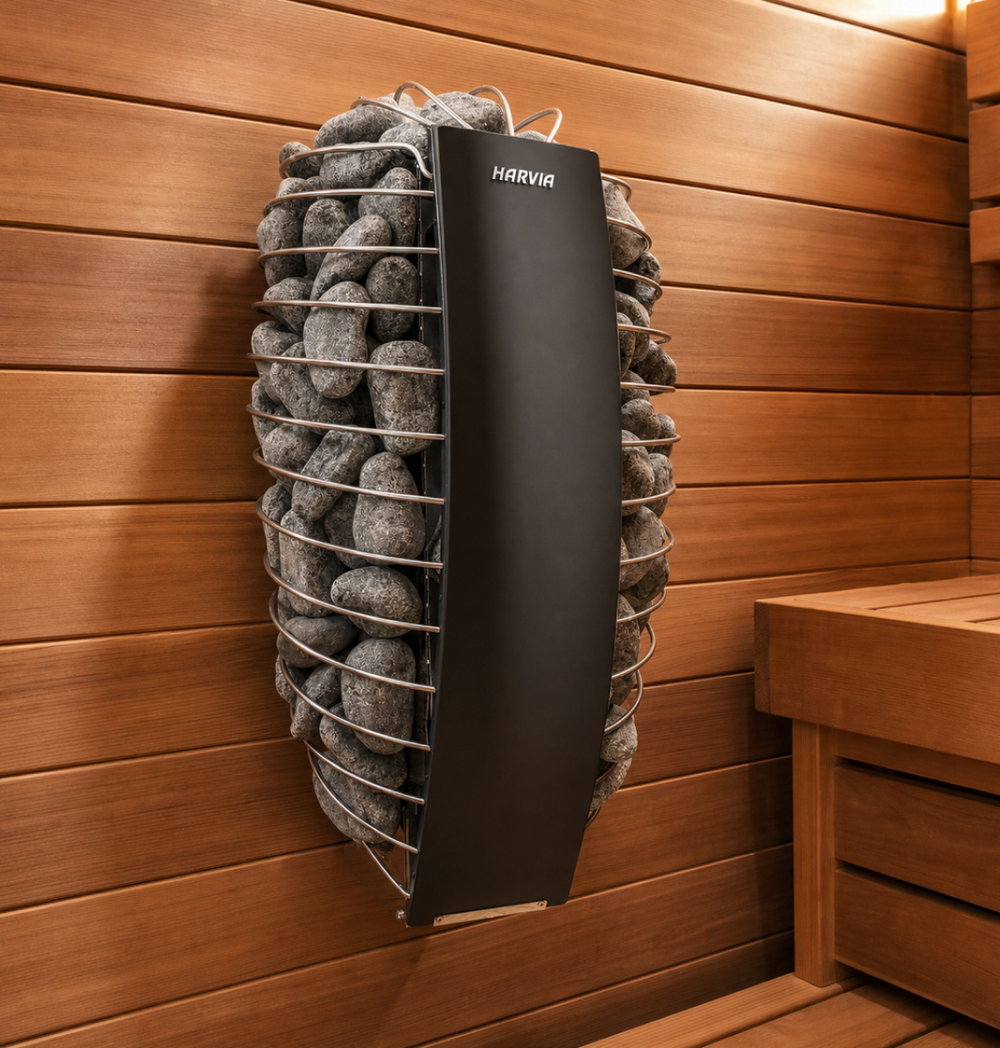 The Baia Glass Sauna features a modern sauna heater filled with gray stones, mounted on a wooden wall inside an indoor sauna room with light brown wood panels and a nearby wooden bench.