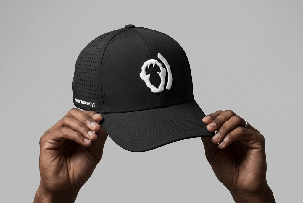A person holds the Polar Monkeys Cap, a black breathable hat with a white embroidered abstract monkey face on the front and "skitmonkeys" text on the side, set against a plain gray background. Part of the Polar Monkeys performance cap collection.