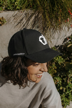 A person with shoulder-length dark hair smiles while wearing a black Polar Monkeys Cap featuring a white monkey logo, sitting outdoors near greenery and sunlight.