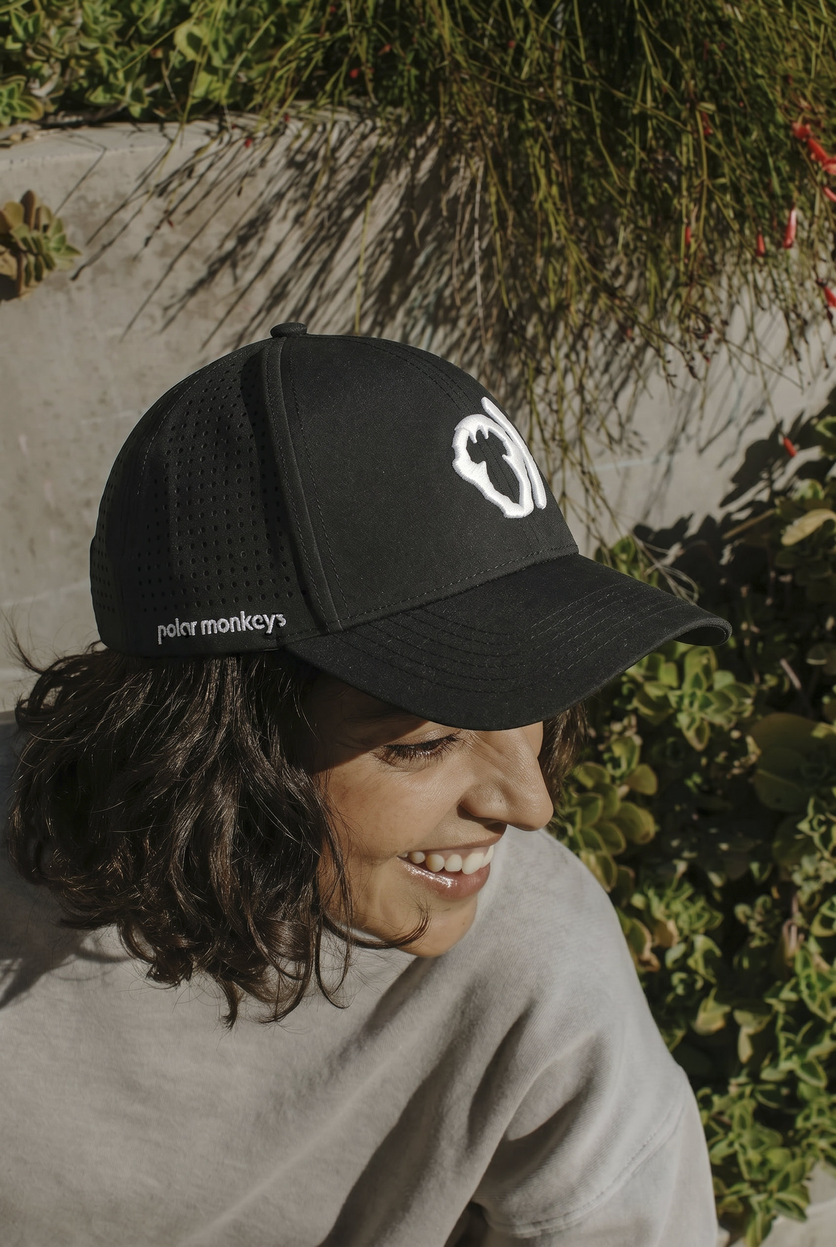A person with shoulder-length dark hair smiles while wearing a black Polar Monkeys Cap featuring a white monkey logo, sitting outdoors near greenery and sunlight.