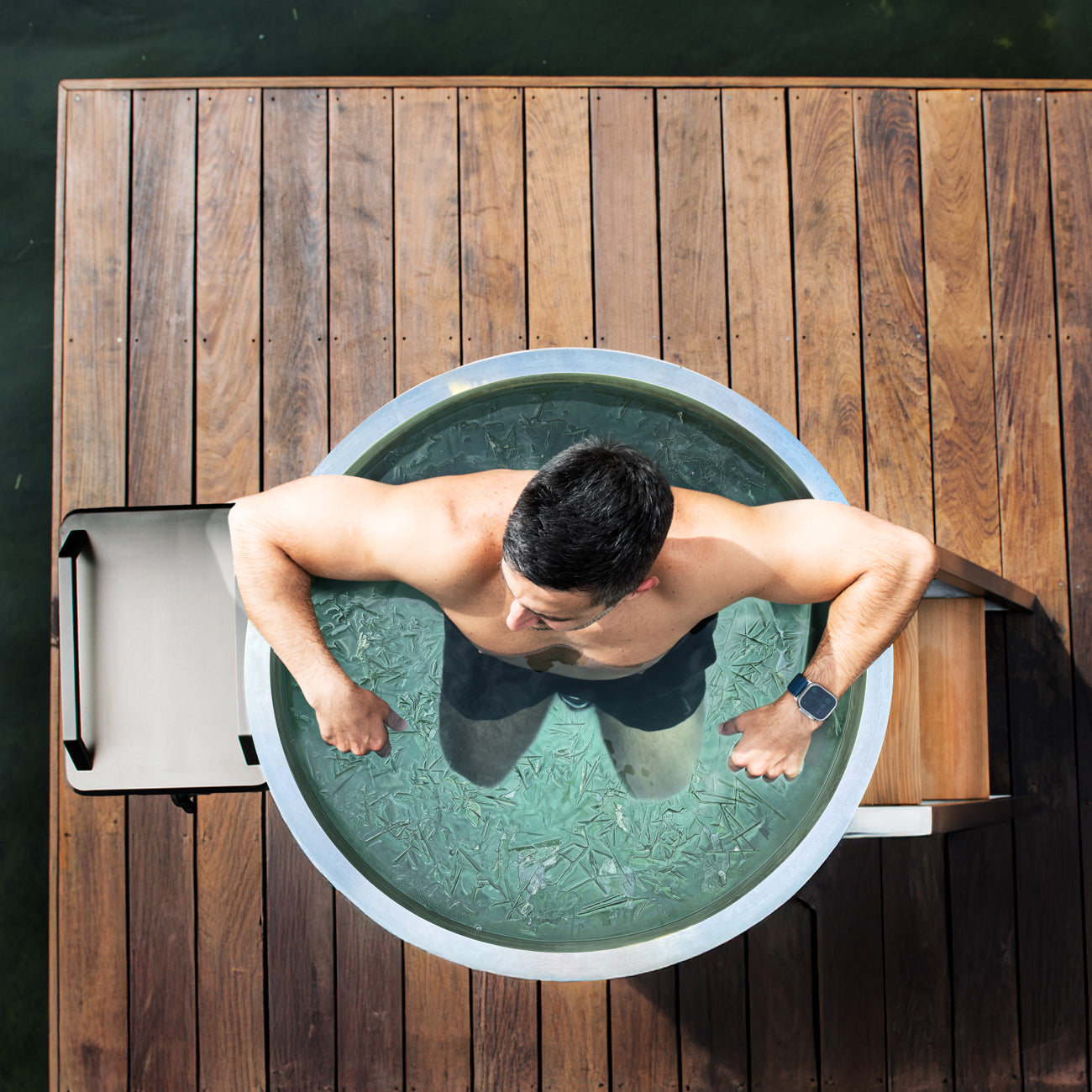 A man relaxes in a Cyber Barrel ice bath on a wooden dock, arms on the edge. He wears a smartwatch and gazes at the icy water inside the vertical marine-grade stainless steel design.