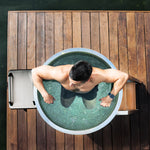 A man relaxes in a Cyber Barrel ice bath on a wooden dock, arms on the edge. He wears a smartwatch and gazes at the icy water inside the vertical marine-grade stainless steel design.