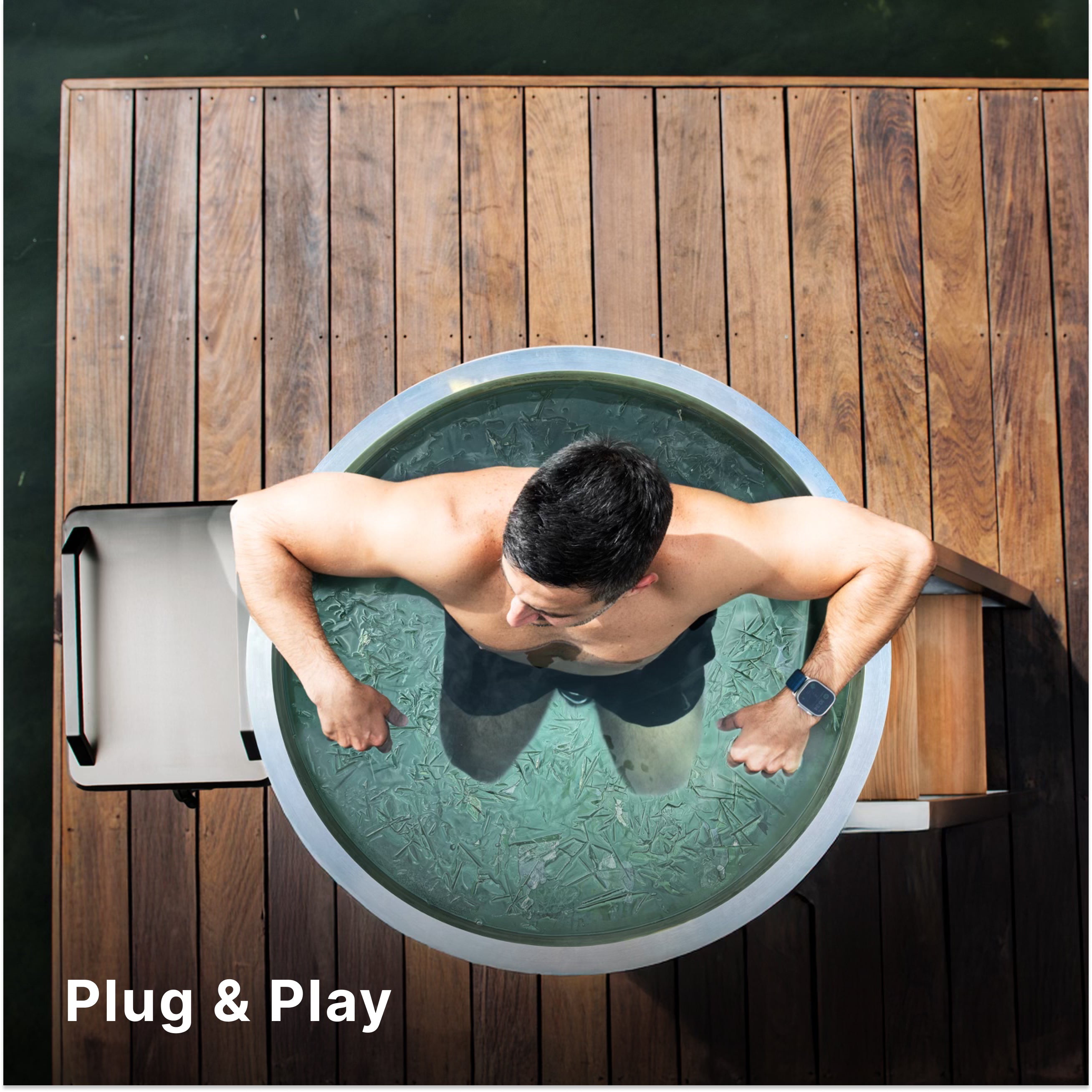 A man sits in a circular ice bath on a wooden dock beside water, viewed from above. He is shirtless and wearing a smartwatch. The text Plug & Play appears in the lower left corner.