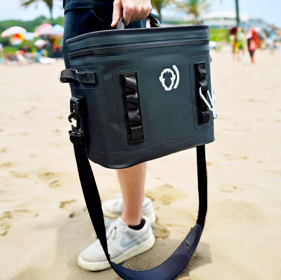 On a sandy beach, someone holds The Cave Cooler—a dark gray soft cooler with cold retention, a shoulder strap, and a white paw print logo—while palm trees and umbrella-covered beachgoers fill the background.