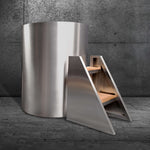 The Cyber Barrel, a modern cylindrical cold plunge crafted from marine-grade stainless steel, rests on a stone floor beside an angular metal and wood step stool, all set against a textured dark concrete wall.
