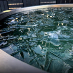 A close-up of the ChillX Chiller, a portable round metal container with green-tinted water and jagged ice pieces for faster cooling cold plunge sessions, reflecting outdoor light and its surroundings.