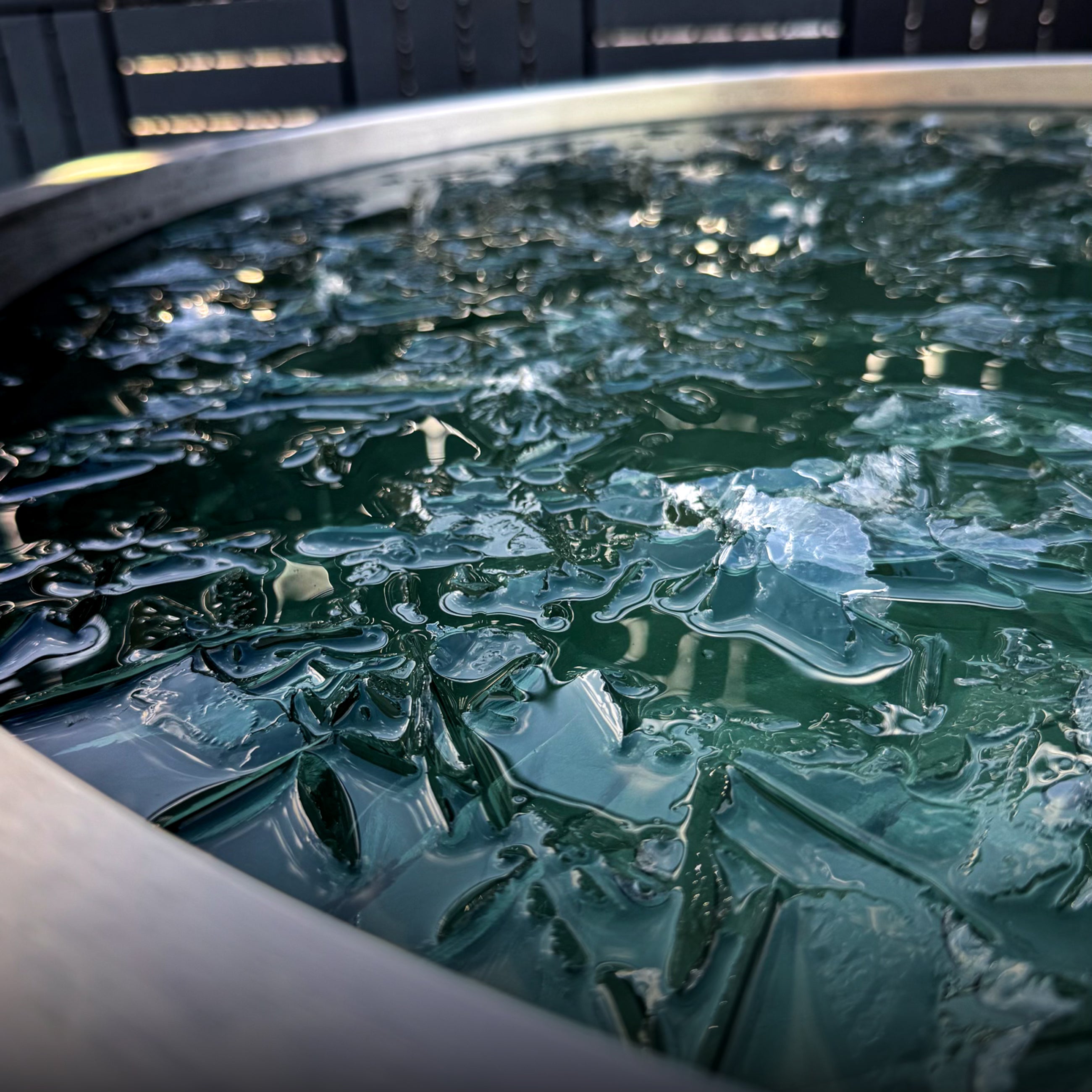 A close-up of the ChillX Chiller, a portable round metal container with green-tinted water and jagged ice pieces for faster cooling cold plunge sessions, reflecting outdoor light and its surroundings.