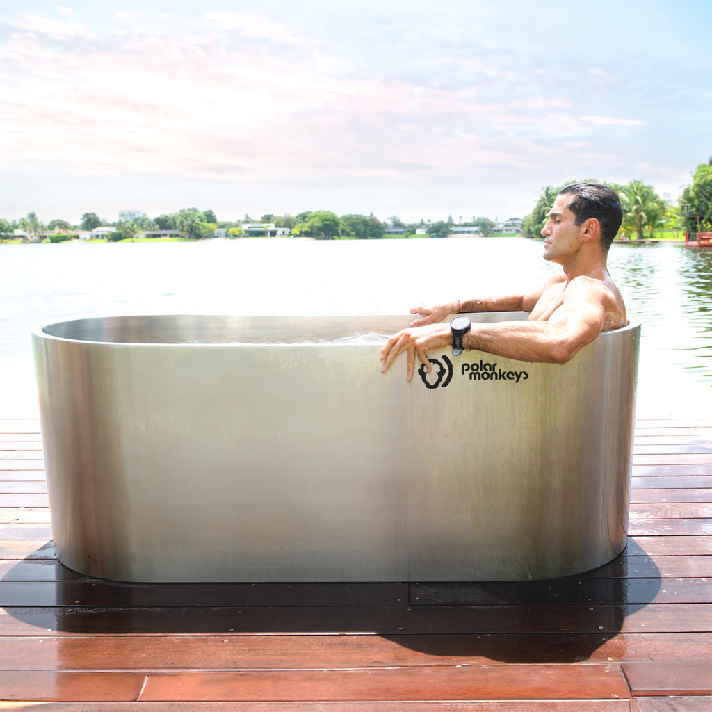 A man relaxes in a Cyber Plunge Commercial Cold Plunge with stainless steel and commercial-grade filtration, on a wooden deck by a lake, surrounded by trees and a pink-blue sky.