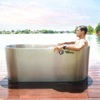 A man relaxes in a Cyber Plunge Commercial Cold Plunge with stainless steel and commercial-grade filtration, on a wooden deck by a lake, surrounded by trees and a pink-blue sky.