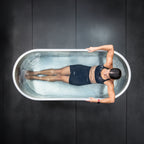 A woman in athletic wear lies in a stainless steel tub filled with water, holding the edges. The dark, minimalistic background emphasizes the Cyber Plunge Commercial Cold Plunge experience.