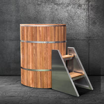 The Star Treatment 2.0 Commercial Cold Plunge, a cylindrical wooden tub with metal bands, rests on a gray stone floor beside a modern metal staircase with wooden steps and a textured gray wall backdrop.