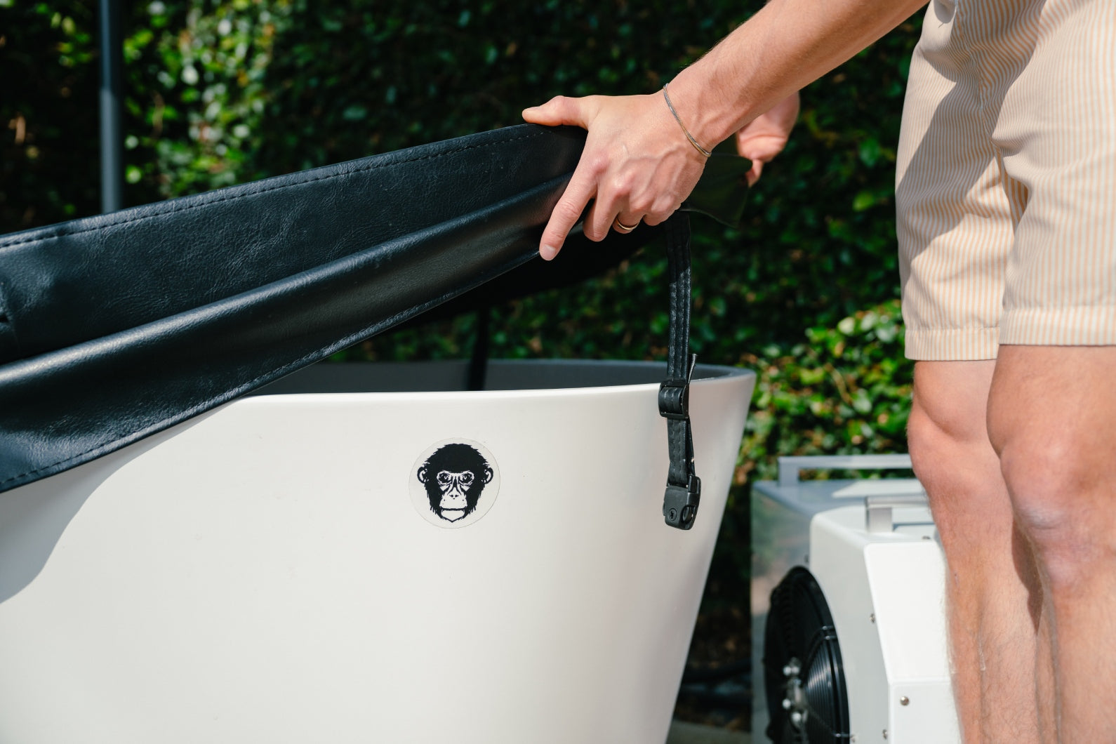 Wearing light shorts, a person lifts the Insulated Cold Plunge Cover—crafted with marine-grade synthetic leather and insulation foam—from a white tub with a monkey face logo, outdoors amid lush greenery.