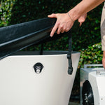 Wearing light shorts, a person lifts the Insulated Cold Plunge Cover—crafted with marine-grade synthetic leather and insulation foam—from a white tub with a monkey face logo, outdoors amid lush greenery.