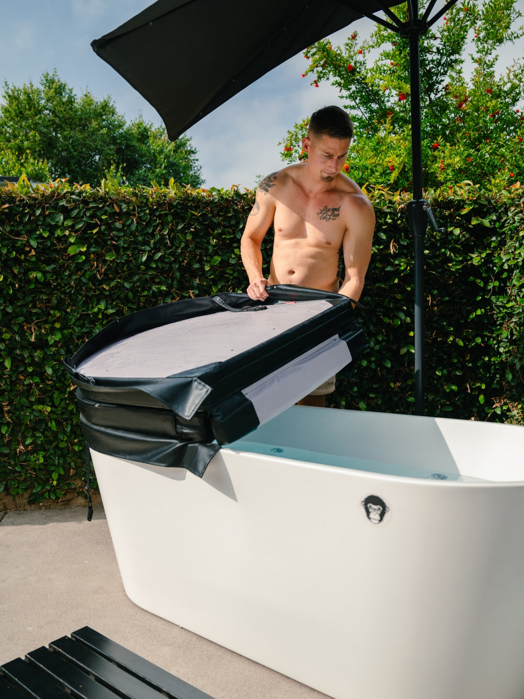 A shirtless man outdoors lifts the Insulated Cold Plunge Cover, crafted from marine-grade synthetic leather, off a white freestanding tub under an umbrella, with green hedges and trees in the background.