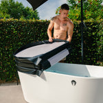 A shirtless man outdoors lifts the Insulated Cold Plunge Cover, crafted from marine-grade synthetic leather, off a white freestanding tub under an umbrella, with green hedges and trees in the background.