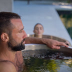 A man with short hair and a beard relaxes in an outdoor tub from the Contrast Edition hot and cold therapy system, while a woman soaks nearby. Both look calm and at ease in this tranquil recovery space.
