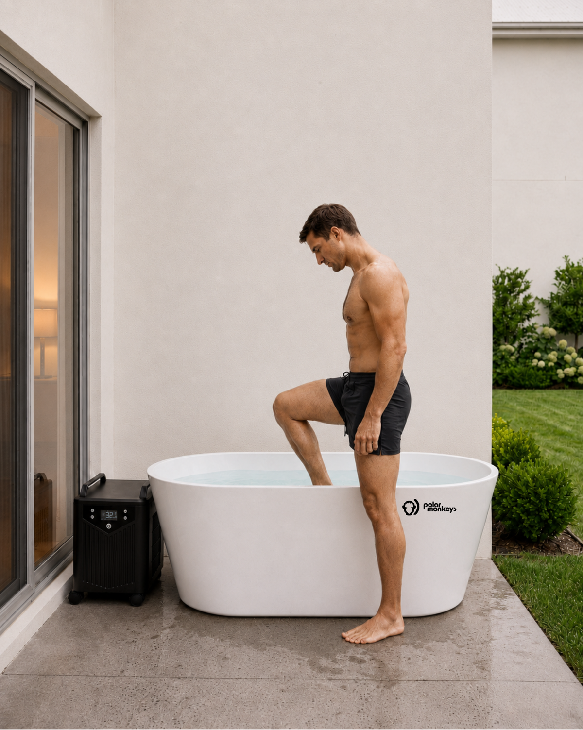 A man in black shorts is stepping into a ONE by Polar Monkeys white outdoor ice bath tub for a cold plunge on a patio next to a house, with greenery and a small black cooling chiller nearby.