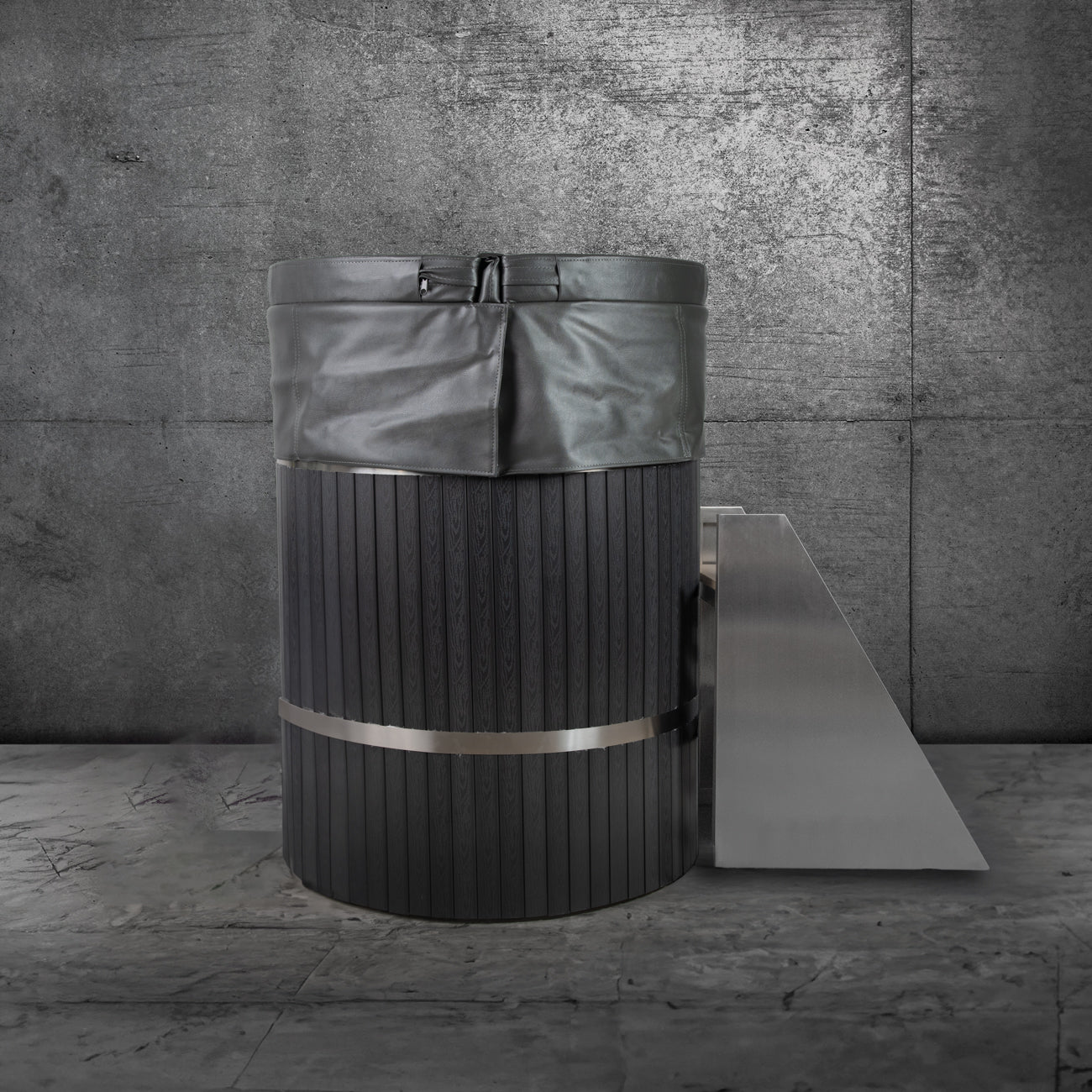 A modern black ice bath with the Insulated Cold Plunge Cover made from marine-grade synthetic leather stands on a stone floor by a concrete wall, featuring a metallic step for easy access.