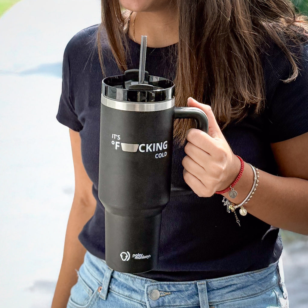 A woman in a navy t-shirt and jeans holds a large Polar Monkeys Tumbler with a straw. This stainless steel tumbler features a funny, partially censored cold-themed phrase—ideal for staying hydrated anywhere.