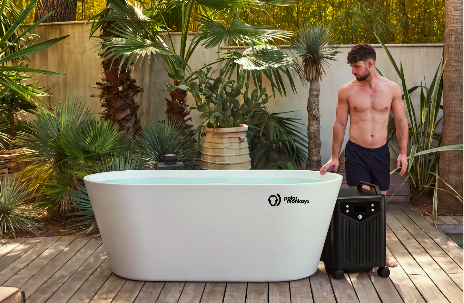 A man in swim trunks stands on a wooden deck beside a white outdoor tub filled with water, surrounded by tropical plants. He touches a black device next to the tub labeled ONE by Polar Monkeys, renowned for its cold plunge setup and advanced ice-making capabilities.
