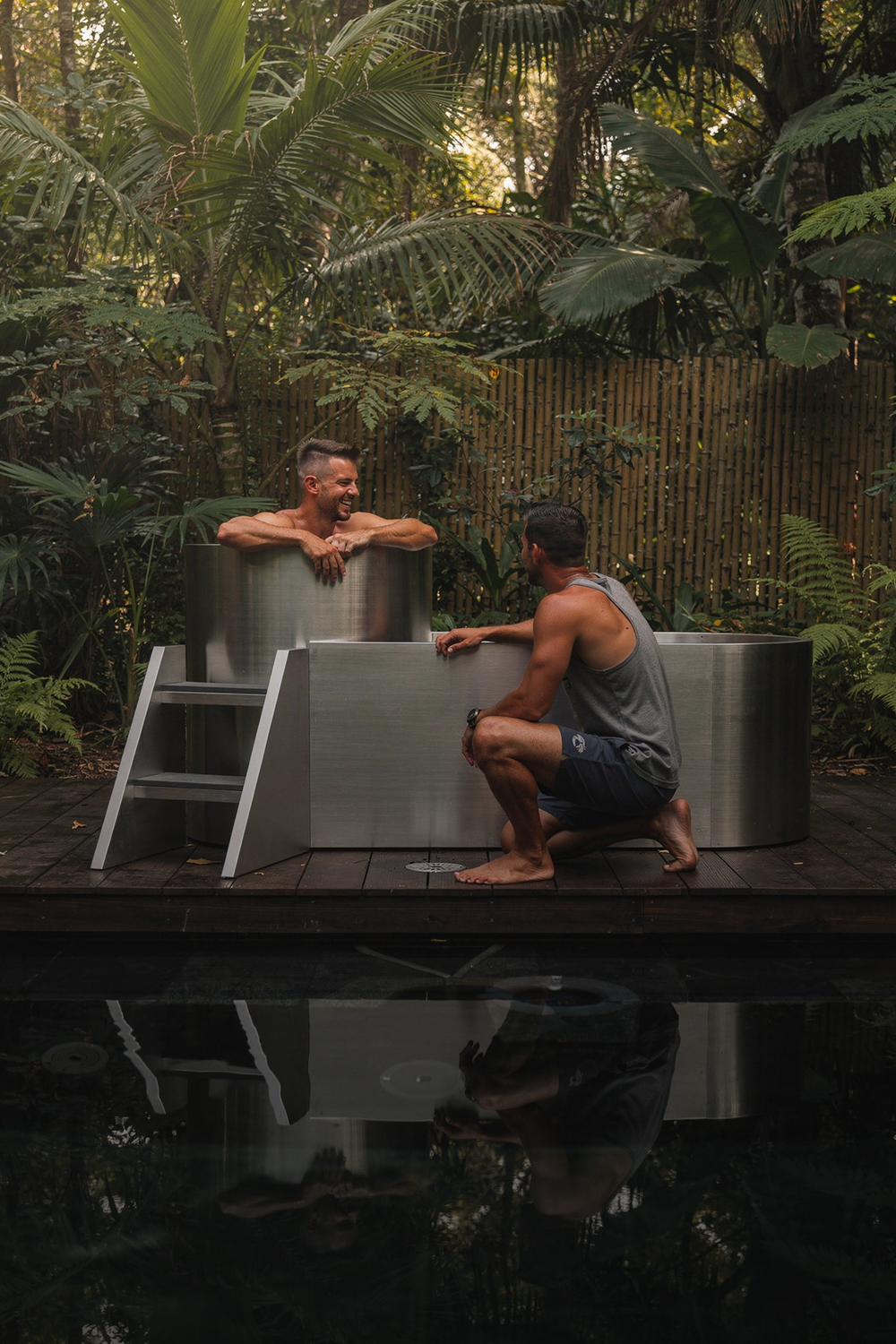 Two men chat in a lush outdoor setting; one sits in the Contrast Edition tub as part of a contrast therapy system, while the other crouches nearby. A white ladder leans on the tub. Greenery and a wooden fence surround them, creating an inviting recovery environment.
