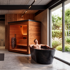 A person enjoys the ultimate home recovery setup for contrast therapy with the Sauna + Cold Plunge Bundle: a sleek black soaking tub is positioned beside a modern wooden sauna in a contemporary room featuring concrete floors and large windows overlooking lush greenery.