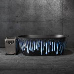 The Brainpod 2.0, a modern black bathtub with blue and white drips and ergonomic design, stands beside a metallic portable machine on a textured concrete floor against a dark gray industrial wall.